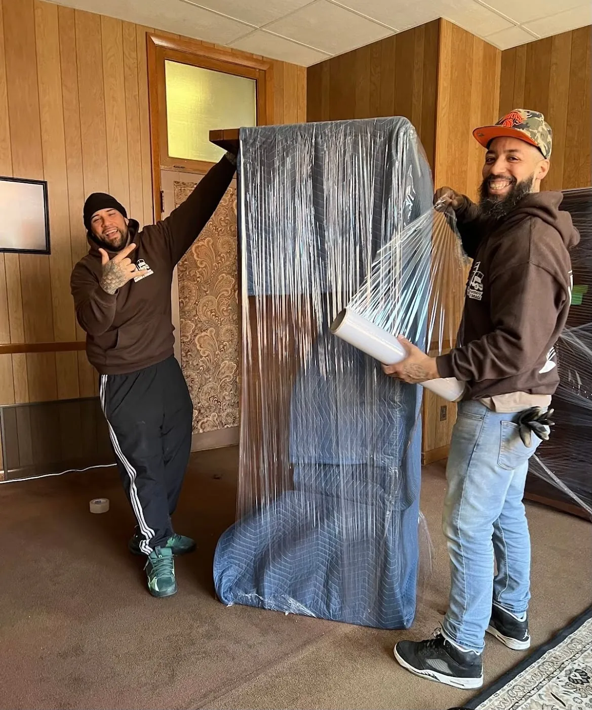 Two movers lifting a wrapped mattress or mirror indoors